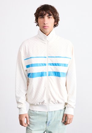 Chaqueta de forro polar en blanco roto con un patrón texturizado. Presenta tres franjas horizontales azules en el pecho y un cuello alto.