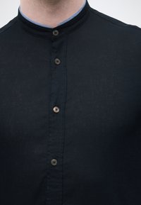 Κοντινό πλάνο ενός ατόμου που φοράει μαύρο πουκάμισο με γιακά μανταρίνι και τέσσερα ορατά κουμπιά, καθώς και γκρι γιακά υποκάμισου.