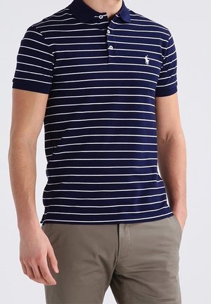 Mann trägt ein marineblaues Polohemd mit weißen horizontalen Streifen und khakifarbene Hose, steht mit einer Hand in der Tasche.