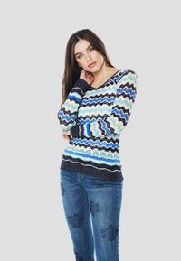Többszínű kötött pulóver hullám mintákkal kék, fehér és fekete színekben, kerek nyakkivágással és hosszú ujjal. Sötétkék farmernadrággal párosítva.