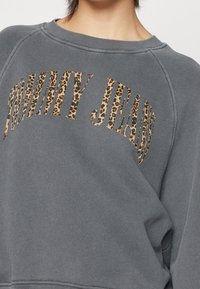 Grå sweatshirt med "TOMMY JEANS" tryckt i leopardmönstrade bokstäver, bärs av en person med kort hår, som slutar precis ovanför kragen.