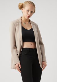 Mladá žena s blond vlasmi na bunách, oblečená v béžovom saku, čiernom crop tope, čiernych džínsoch a farebnom náhrdelníku s kvetmi, na bielom pozadí.