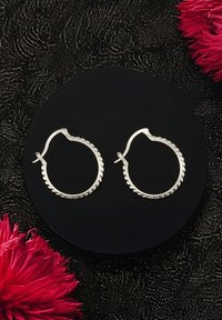 Silvriga kreolörhängen med en texturerad, vriden kant, visade på ett runt svart stativ mot en mörk bakgrund.