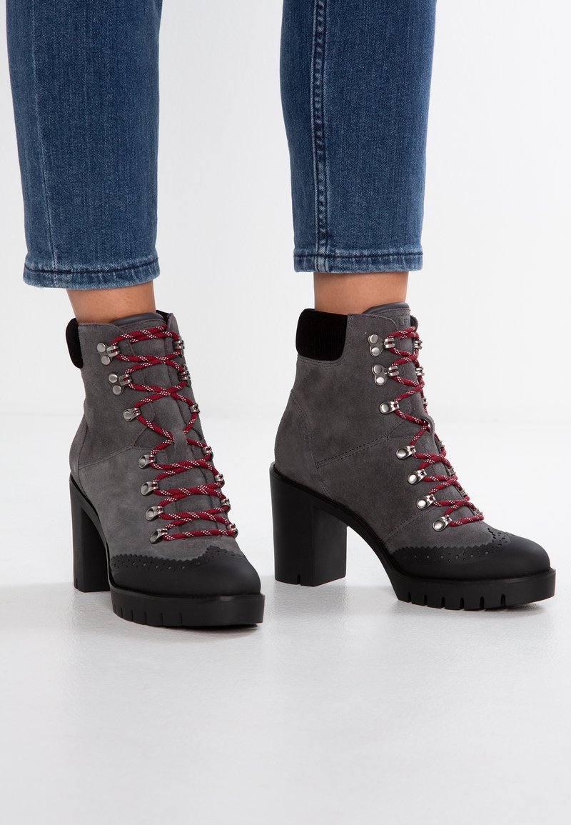 Bottes montantes en daim gris à lacets avec bouts noirs, talons hauts épais, lacets rouges, portées avec un jean bleu raccourci.
