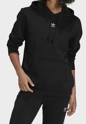 Osoba w czarnej bluzie z kapturem Adidas i dopasowanych czarnych spodniach z białymi logo Adidas na klatce piersiowej i udzie, stoi z rozluźnionymi rękami.