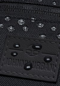 Svart texturerad tyg med upphöjda vattendroppar och en svart läderlapp med präglad "JOHN URBAN" och cirkulär logotypdesign.