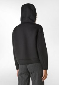 Felpa nera con cappuccio, dal taglio rilassato, orlo a coste e texture morbida, abbinata a pantaloni grigi con righe sottili. Disegno minimalista in evidenza.
