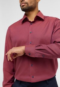 Camicia rossa a fantasia con maniche lunghe, caratterizzata da un colletto con bottoni e bottoni blu, indossata con una cintura scura.