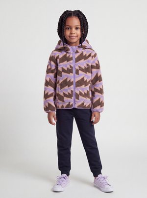 Jeune fille avec des cheveux tressés portant un sweat à capuche zippé marron et violet à motifs, un pantalon bleu marine et des baskets violet clair, se tenant devant un fond uni.