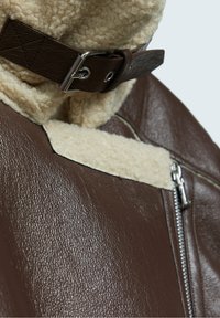 Brązowa skórzana kurtka z miękkim, kremowym kołnierzem z kożucha, metalowym zamkiem błyskawicznym i wyraźnym zapięciem na kołnierzu.