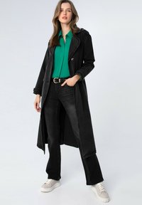Cappotto lungo nero con colletto a risvolto, design doppiopetto, abbinato a una camicia verde, jeans neri e sneakers grigio chiaro.