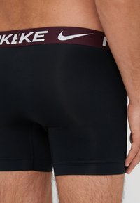 Čierne boxerky s bordovým elastickým pásom a bielym logom Nike. Materiál vyzerá hladko a má priliehavý strih.