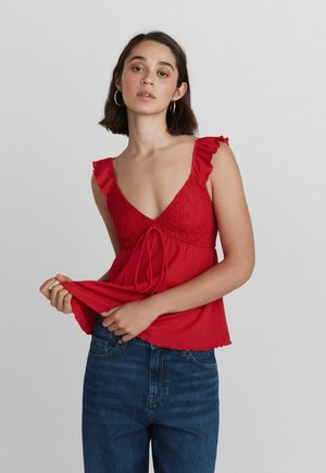 Jeune femme portant un haut rouge sans manches avec des volants et un jean bleu, tenant l'ourlet de son haut sur un fond clair uni.