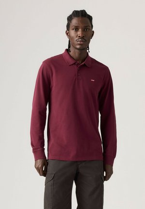 Mand med flettet hår iført en langærmet bordeaux polo og mørke cargobukser mod en ensfarvet lys baggrund.