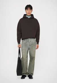 Felpa con cappuccio marrone scuro con logo in rilievo, abbinata a jeans grigi chiari con macchie. Scarpe nere e una borsa a tracolla nera.
