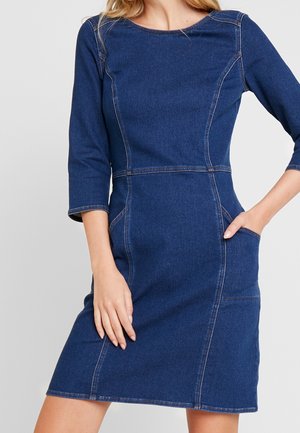 Kobieta w niebieskiej sukience z denimu o długości do kolan, z rękawami do połowy ramienia i kieszeniami, pozująca z jedną ręką w kieszeni.