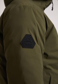 Olivengrøn jakke med et tekstureret sort hexagonalt patch med "JACKIT" præget, synlig syning og en mat finish.