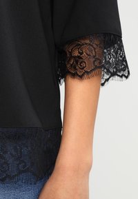 Top nero con maniche corte, caratterizzato da un intricato bordo in pizzo nero all'orlo e alle maniche, che mette in evidenza una texture di tessuto liscia.