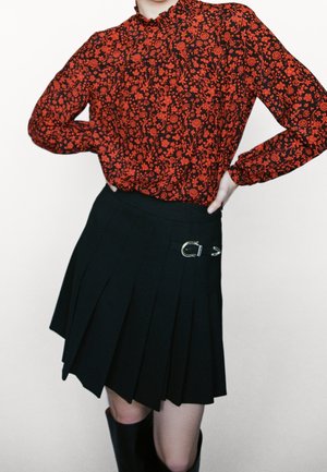 Person trägt eine langärmelige rote Blumenbluse, in einen schwarzen plissierten Rock mit seitlicher silberner Schnalle gesteckt, Hände in der Hüfte.