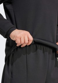 Black athletic outfit featuring a relaxed fit. The top has long sleeves, while the pants exhibit a gathered waistband and side zipper pockets.