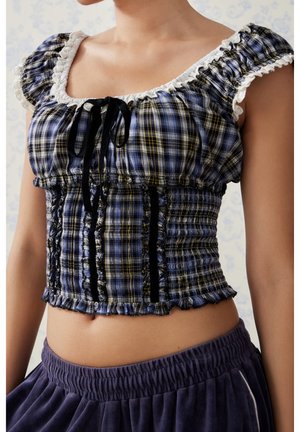 Kobieta ubrana w niebiesko-żółty kraciasty marszczony krótki top z białą falbanką i czarnymi wstążkami, w połączeniu z ciemnymi spodniami z elastycznym pasem.