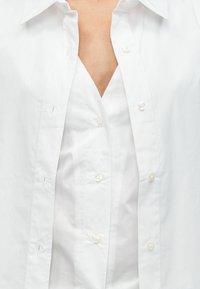 Κοντινό πλάνο ενός ατόμου που φοράει λευκό πουκάμισο με κουμπιά πάνω από λευκό τοπ με V-λαιμόκοψη.