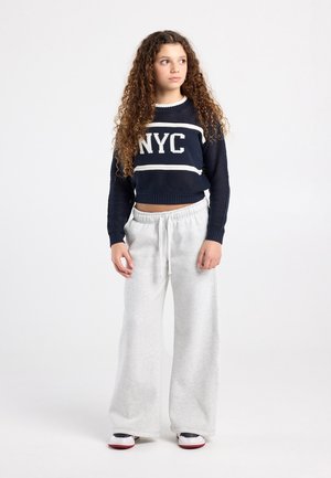 Marineblauwe gebreide trui met tekst "NYC", gecombineerd met lichtgrijze sweatpants met wijde pijpen en witte sneakers. De trui heeft geribbelde boorden en zoom.