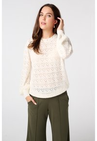 Pull en tricot couleur crème avec des motifs en forme de cœur, col rond et manches longues. Associé à un pantalon large olive.