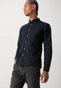 Camisa azul oscuro de botones con mangas largas, corte ajustado y pequeños botones blancos, combinada con pantalones de mezclilla grises. Textura suave, estilo de cuello.