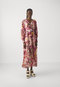 Floral dress in roze en paars, met pofmouwen, ruches en een V-hals. Het materiaal lijkt licht en vloeiend. Draagt met witte platformschoenen.