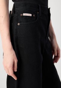 Juodos džinsinio audinio kelnės su matomu raudonu Calvin Klein logotipu, sidabrinėmis detalėmis ir tiesia kojų siluetu. Audinys turi šiek tiek tekstūrinį paviršių.