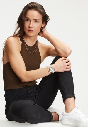 Braun gerippter Haltertop, schwarze eng anliegende Jeans, silberne Uhr mit rundem Zifferblatt, gestapelte Halsketten und weiße Sneakers. Glatte Texturen und schlichte Gestaltung.