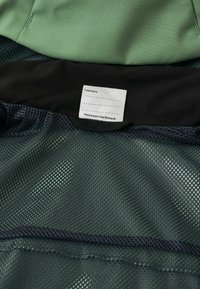 Grüner Stoff, der schwarzen Netzstoff überlagert, mit einem weißen Etikett zur Personalisierung mit dem Text: "Ich war hier: _____ Bitte gib mich weiter!"