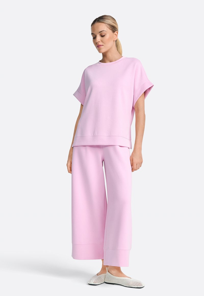 Mujer vestida con una blusa holgada de manga corta en color rosa claro y pantalones de pierna ancha, con zapatos tipo slip-on tejidos de color crema, de pie sobre un fondo blanco liso.