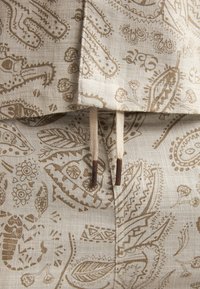 Tissu beige avec des motifs cachemire marron complexes, doté d'un cordon de serrage avec des accents en bois à la taille. Matériau texturé et léger.