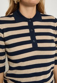 Strikket polo med marineblå og beige horisontale striber, med krave og tre-knaps lukning, tekstureret materiale.