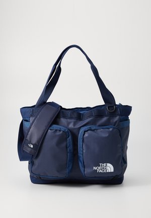 Sac fourre-tout bleu marine en tissu durable, avec deux poches avant, une bandoulière, des poignées supérieures et un détail de logo blanc.