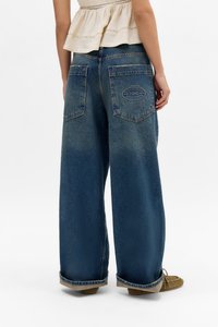 Person som har på seg vide blå denimjeans med oppbrettede ankler og baklomme, kombinert med brune mokkasinsko.