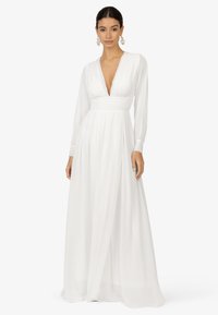Robe blanche à manches longues avec un décolleté en V profond, taille empire et jupe fluide. Tissu doux créant une texture lisse ; poignets ajustés avec des détails de boutons.