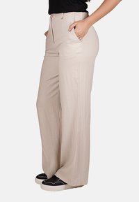 Pantaloni beige a gamba larga realizzati in un tessuto leggero, con tasche laterali, zip frontale e un design a vita alta. Indossati con sneakers nere.