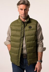 Olivově zelená prošívaná vesta s předním zipem, horizontálním prošitím a logem. Nosená přes béžovou kostkovanou košili a tmavé džíny.