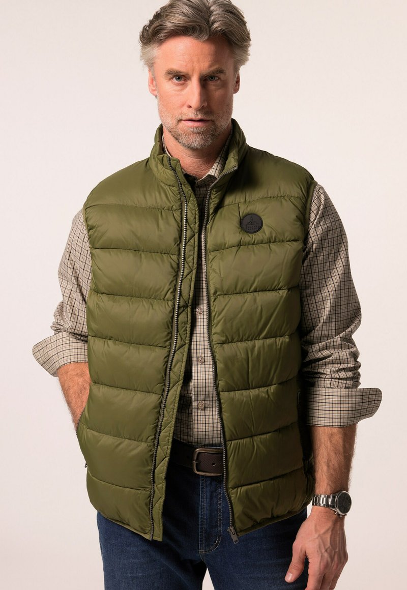 Olivově zelená prošívaná vesta s předním zipem, horizontálním prošitím a logem. Nosená přes béžovou kostkovanou košili a tmavé džíny.