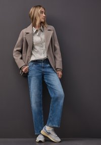 Beżowy płaszcz wełniany, jasnoniebieska koszula w paski, wyblakłe niebieskie dżinsy oraz szare sneakersy z żółtymi akcentami, stojący na tle ciemnego tła.