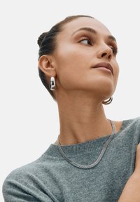 Silverörhängen i ringform och ett texturerat silverkedjehalsband bärs tillsammans med en grå tröja. Smyckena har en jämn, blänkande yta.