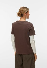 Καφέ κοντομάνικη μπλούζα από μαλακό ύφασμα με στρογγυλή λαιμόκοψη, φορεμένη πάνω από λευκό μακρυμάνικο πουκάμισο, εμφανιζόμενη από πίσω.