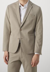 Uomo che indossa una giacca beige coordinata e pantaloni abbinati sopra una camicia bianca a maniche lunghe, in piedi davanti a uno sfondo chiaro e uniforme.