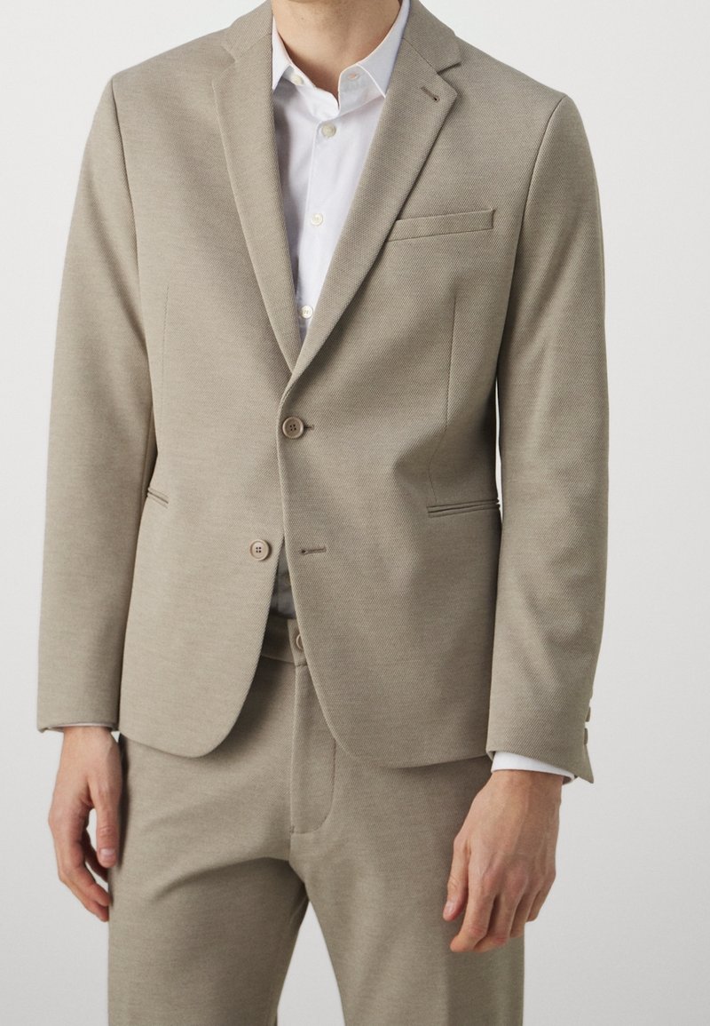 Uomo che indossa una giacca beige coordinata e pantaloni abbinati sopra una camicia bianca a maniche lunghe, in piedi davanti a uno sfondo chiaro e uniforme.