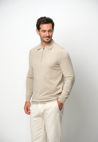 Beige Strickpullover mit langen Ärmeln, Reißverschlusskragen und Rippbündchen, getragen mit hellen Hosen. Weicher Stoff und figurbetontes Design.