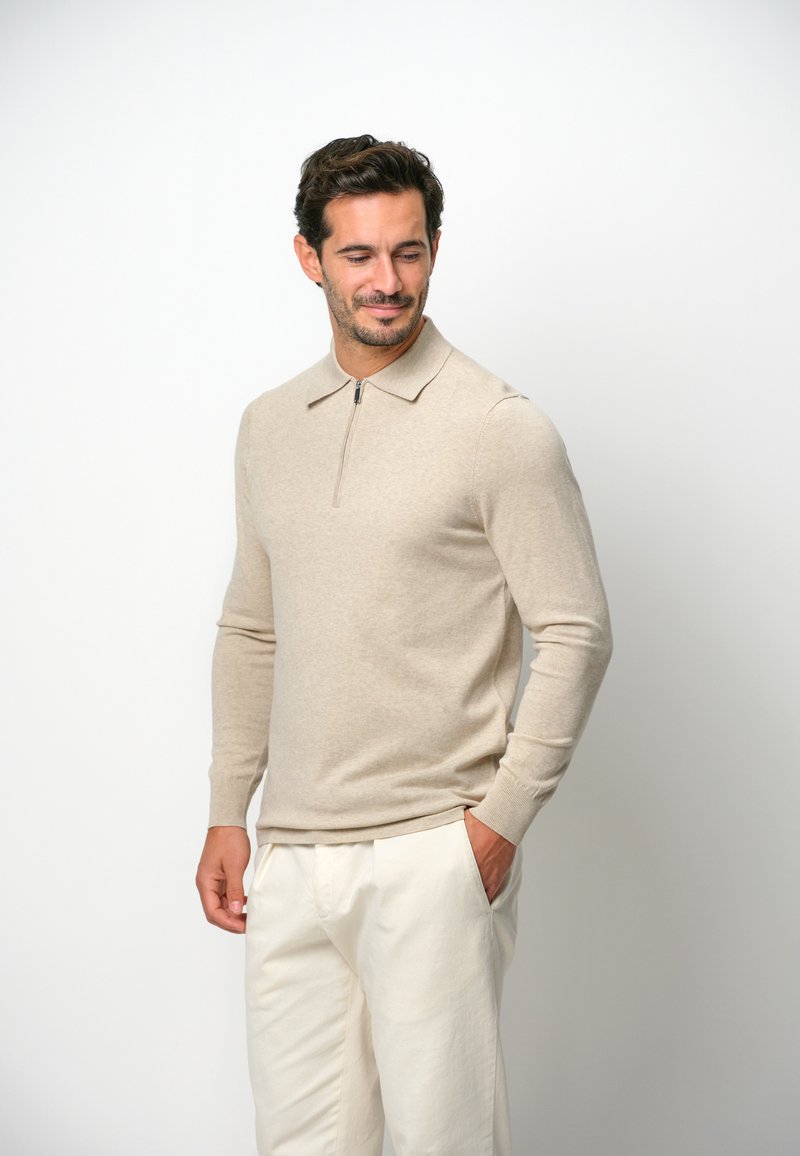 Beige Strickpullover mit langen Ärmeln, Reißverschlusskragen und Rippbündchen, getragen mit hellen Hosen. Weicher Stoff und figurbetontes Design.