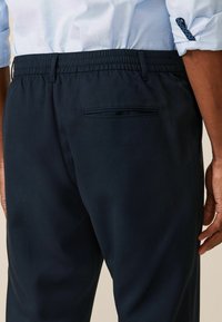 Marineblaue Hose mit elastischem Bund, glatter Textur und einer Rückentasche mit einfachem Design. Eine teilweise sichtbare Hand an der Seite.